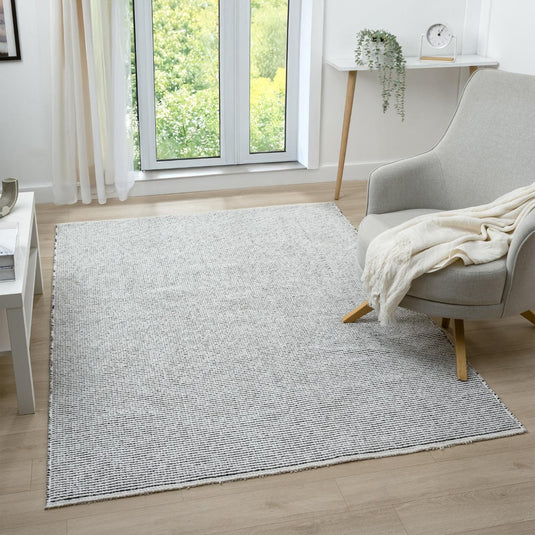 Light gray rug on a wooden floor with a chair and window in the background